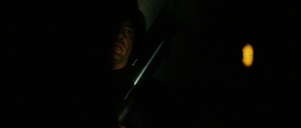 No Country For Old Men movie still: closeup — A man stands in a dark environment with a single faint light source visible. He holds a dark object…