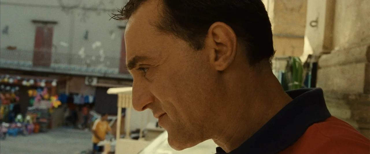 Reality (2023) movie still: closeup — An outdoor balcony overlooks a blurred sunlit urban plaza with small vendor stalls. A middle-aged man in a…