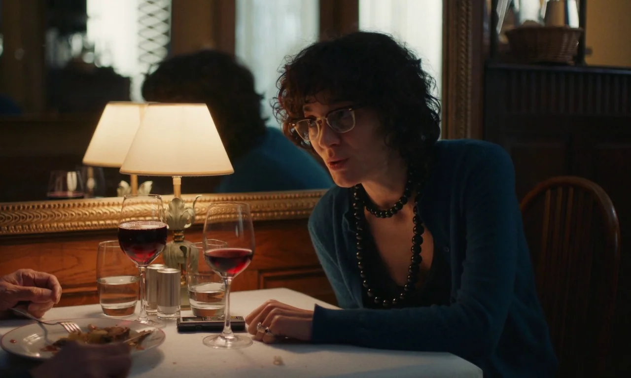 Little Girl Blue (2023) movie still: medium — A dimly lit table at a restaurant features a small lamp and two wine glasses. A woman with dark curly…
