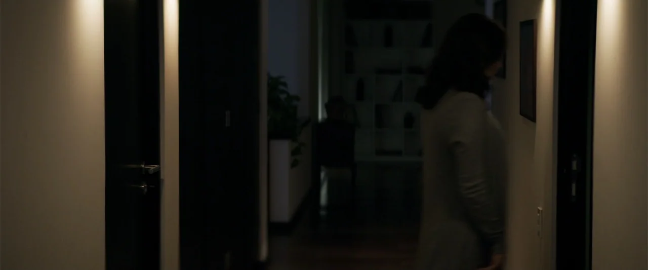 Human Animals (2020) movie still: medium — A narrow residential hallway features white walls, dark doors, and warm wall sconces. A woman in a gray…