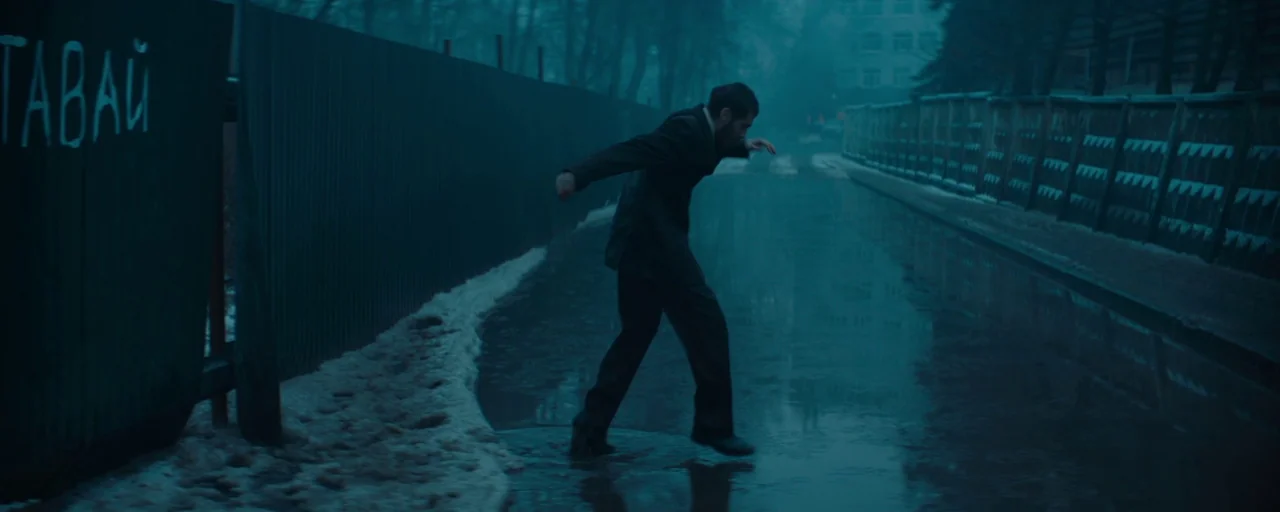 Petrov’s Flu (2021) movie still: fullbody — A narrow outdoor passage bordered by a dark corrugated metal fence with white lettering. A man in a dark…