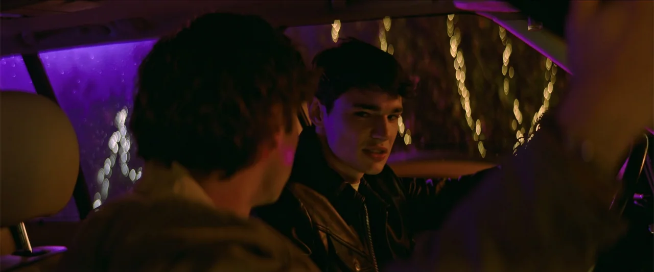 Tell Me Softly (2025) movie still: medium — The interior of a car at night with purple ambient lighting and gold bokeh. A young man sits in the…
