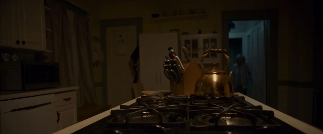 Jump, Darling (2022) movie still: medium — A dimly lit kitchen features a copper tea kettle and knife block on a stove. A silhouette of a woman…