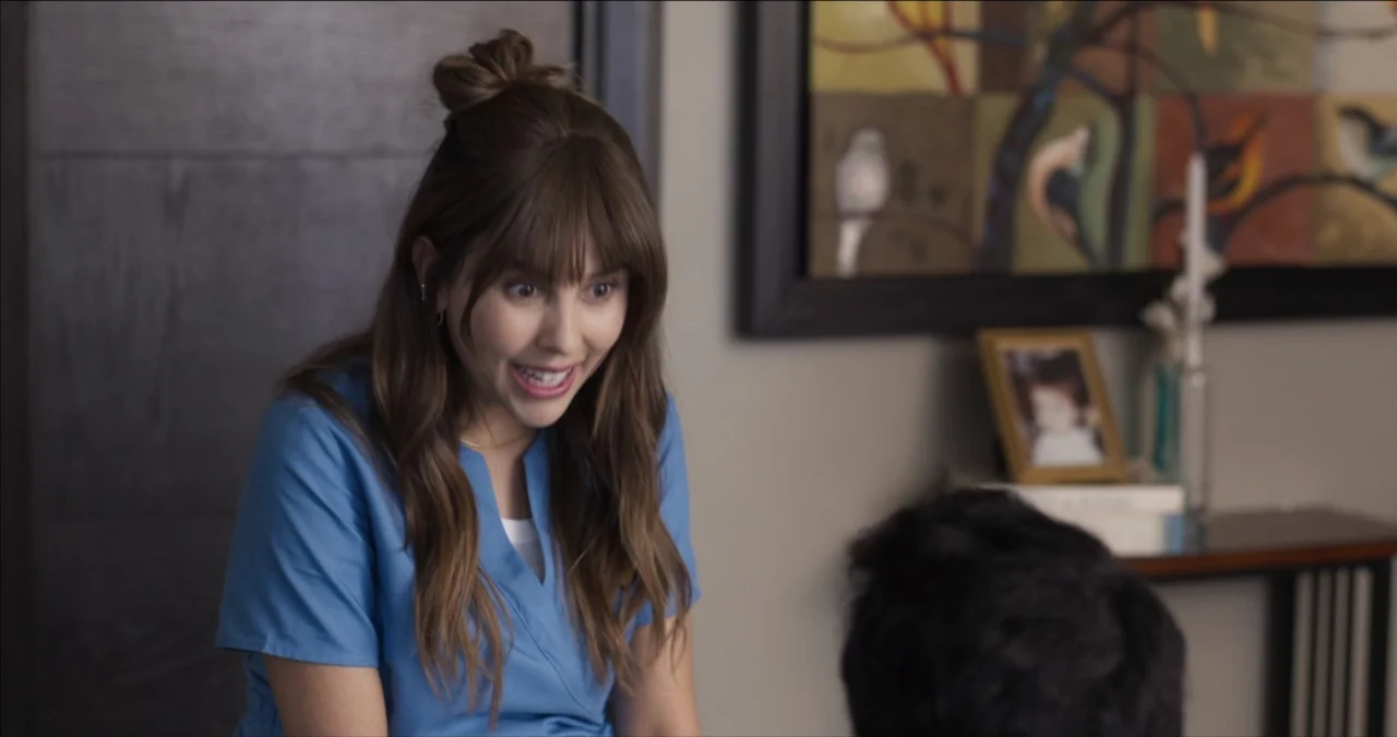 Bendita Suegra (2023) movie still: medium — A residential room features a wall with a framed picture and a console table. A young woman in a blue…