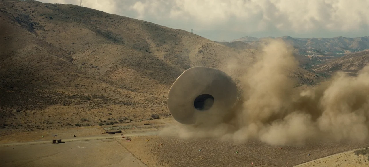 Nope (2022) movie still: wide — A dry, hilly landscape features a fenced-in ranch area under a cloudy sky. A large, smooth, disc-shaped object…