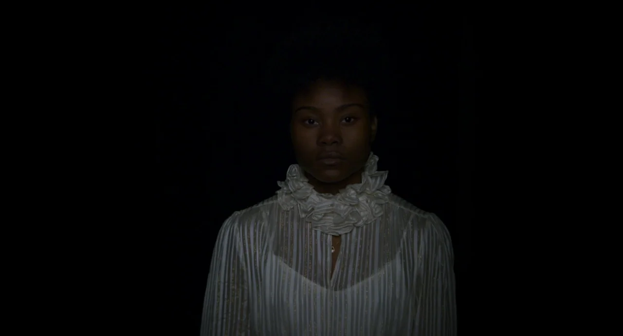 Zombi Child (2019) movie still: medium — A person stands centered against a deep black, undifferentiated background. The subject is wearing a…