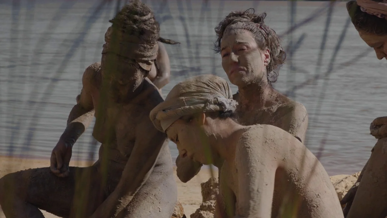 Zama (2017) movie still: medium — A group of women covered in thick mud sit along a sandy riverbank. They remain still in the daylight while their…