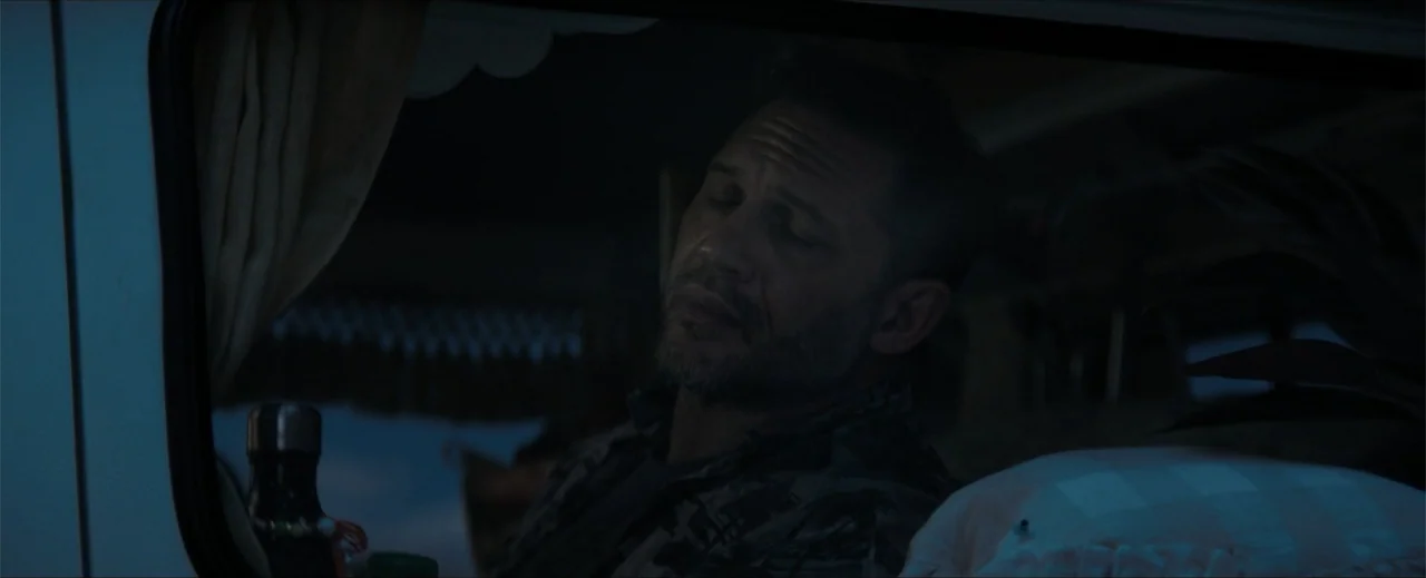 Venom: The Last Dance (2024) movie still: closeup — Interior of a small camper van at night with blue ambient light. A middle-aged man with stubble…