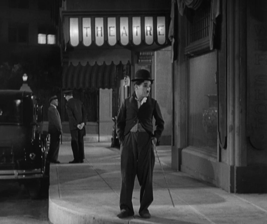 City Lights (1931) movie still: fullbody — A night city sidewalk in front of a theatre with a lit marquee. A man in a bowler hat and oversized suit…