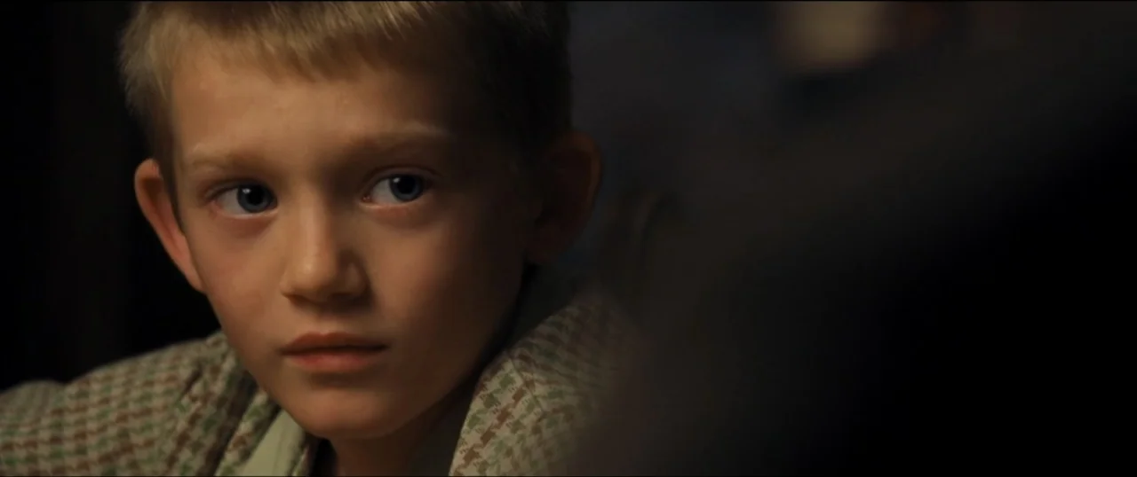 First Man (2018) movie still: closeup — A dimly lit indoor space with an out-of-focus foreground object obscuring part of the view. A young boy with…