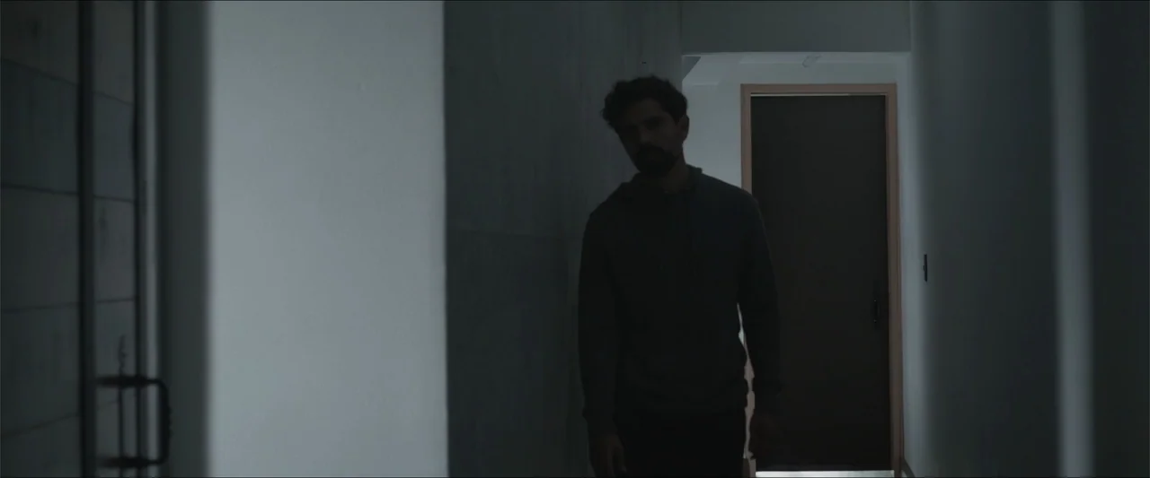 Los habitantes (2023) movie still: medium — A narrow apartment corridor with white walls and a dark door at the end. A man in a grey hoodie walks…