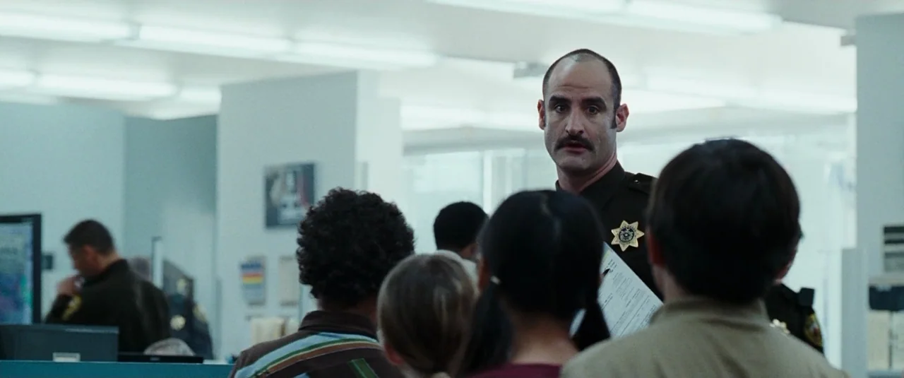 The Hangover (2009) movie still: medium — A sterile government office space with beige walls and drop-ceiling fluorescent lighting. An officer in…