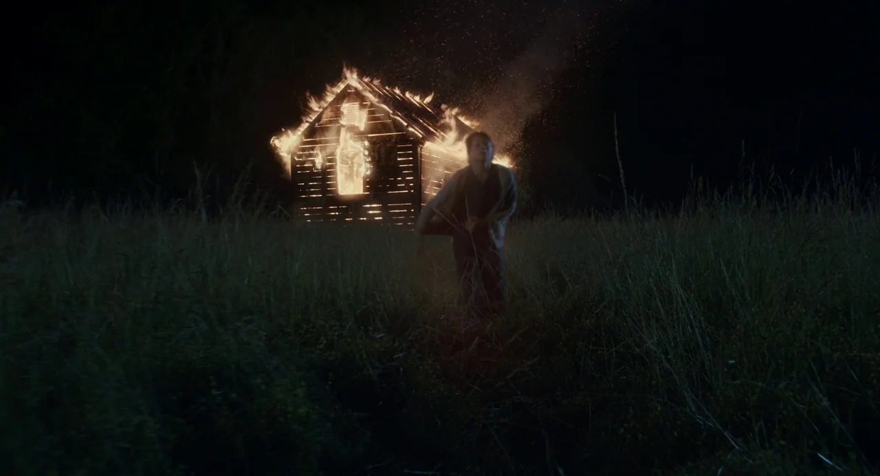 Goodnight Mommy (2022) movie still: wide — A dark night landscape featuring a wooden building engulfed in flames. A young man runs across the tall…