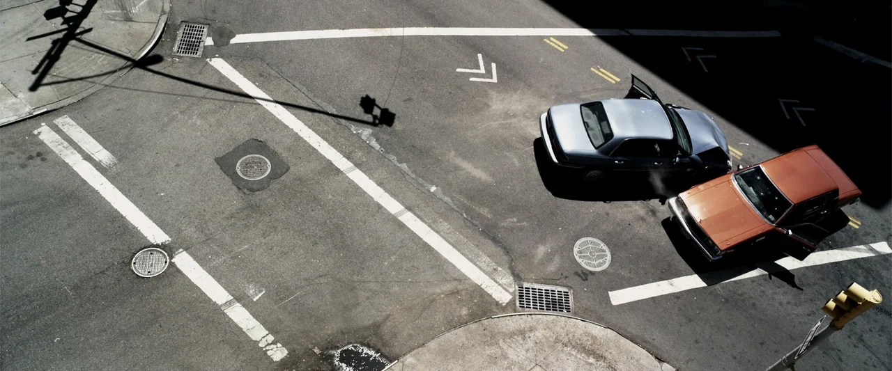 A Walk Among the Tombstones (2014) movie still: wide — An urban asphalt intersection is marked by faded white paint lines and metal manholes. A…
