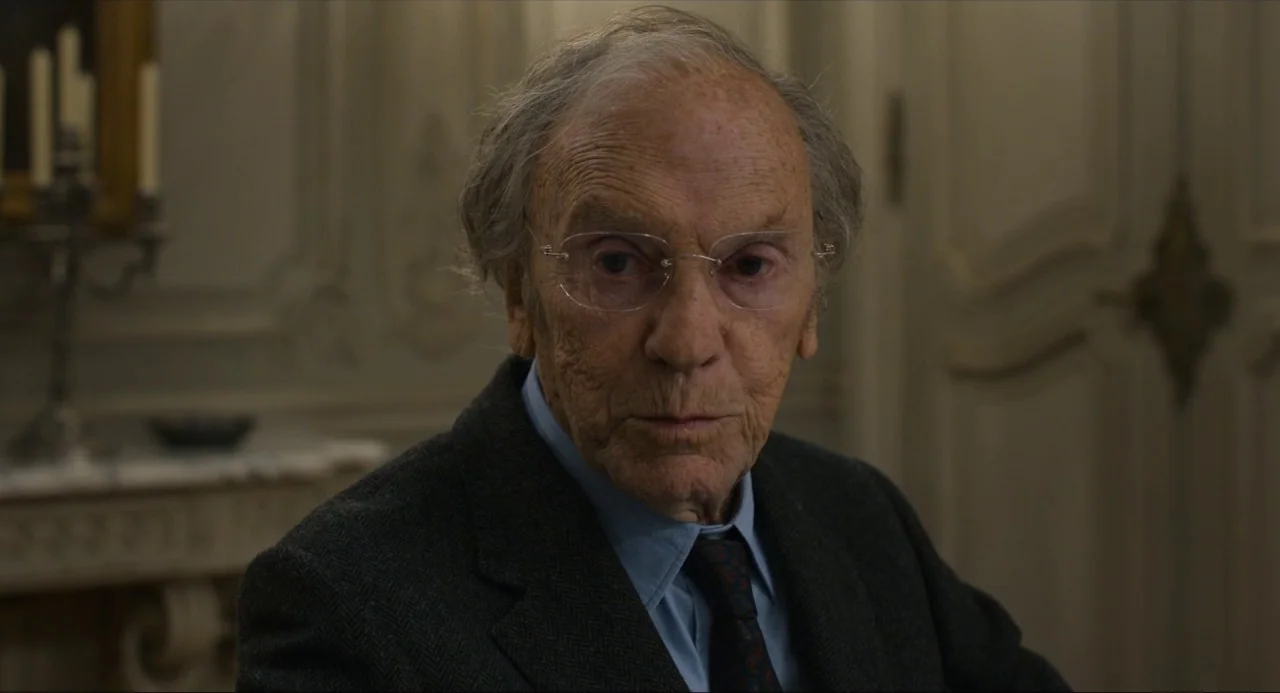 Happy End (2017) movie still: closeup — An elegant drawing room with classic moldings and a marble fireplace in the background. An elderly man in a…