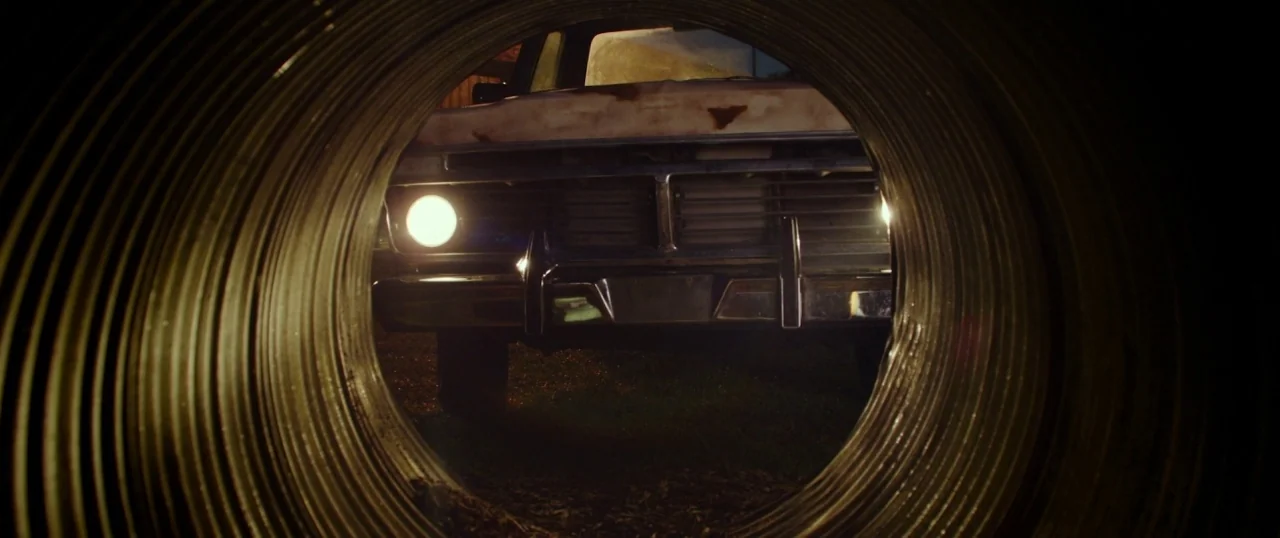 The Strangers: Prey at Night (2018) movie still: wide — A dark, corrugated metal pipe frames a view looking out at a parked vehicle. The front end…