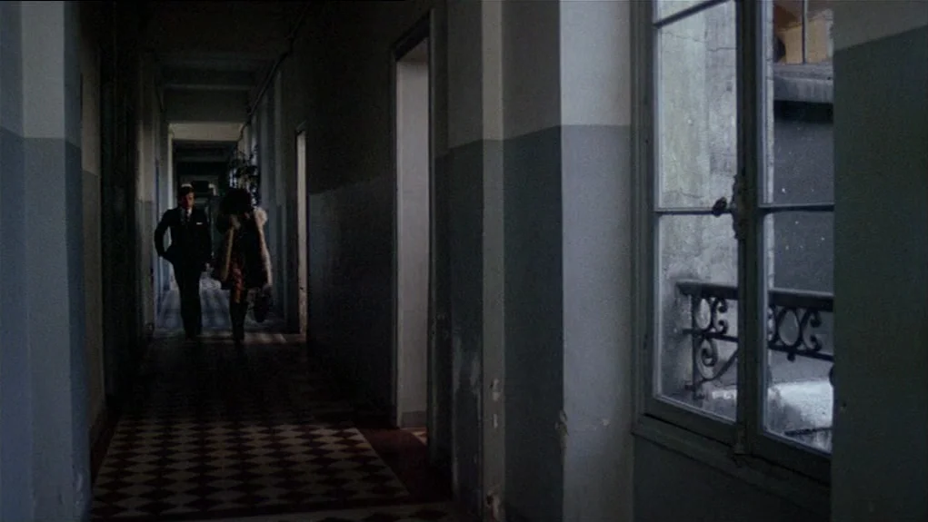 The Tenant (1976) movie still: wide — A long, narrow, dimly lit apartment hallway features a checkered tile floor and windows. A man and a woman…
