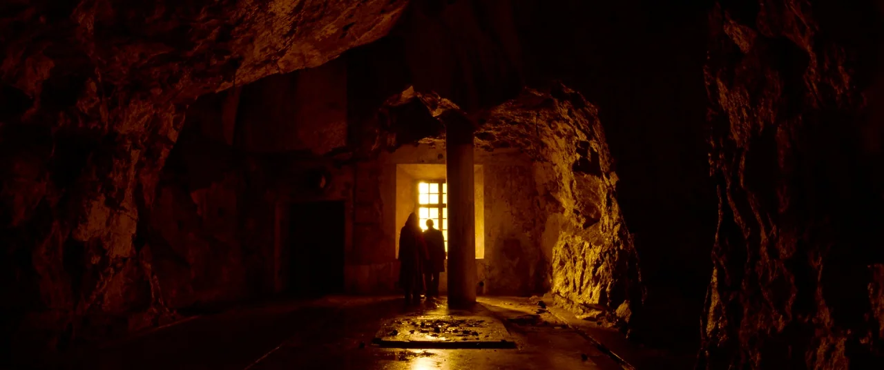The House That Jack Built (2018) movie still: wide — A dark subterranean stone chamber features rough cavernous walls and a single stone pillar. Two…