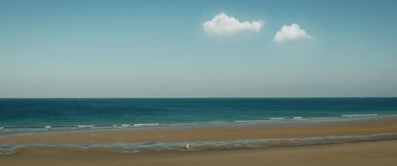Beast (2017) movie still: wide — A vast, expansive beach stretches across the frame with calm blue ocean water. A single small human figure walks…