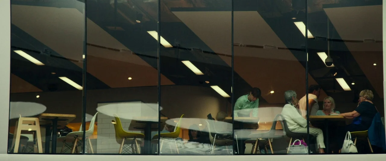 Wild Rose (2018) movie still: wide — A view through a large glass window shows a modern cafe interior with tables. Several people sit at tables…