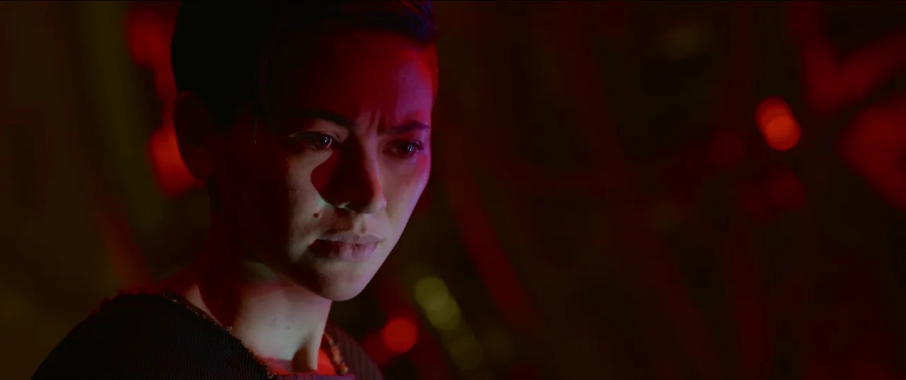 The Matrix Resurrections (2021) movie still: closeup — A dimly lit club interior characterized by deep red ambient light. A young woman with short…