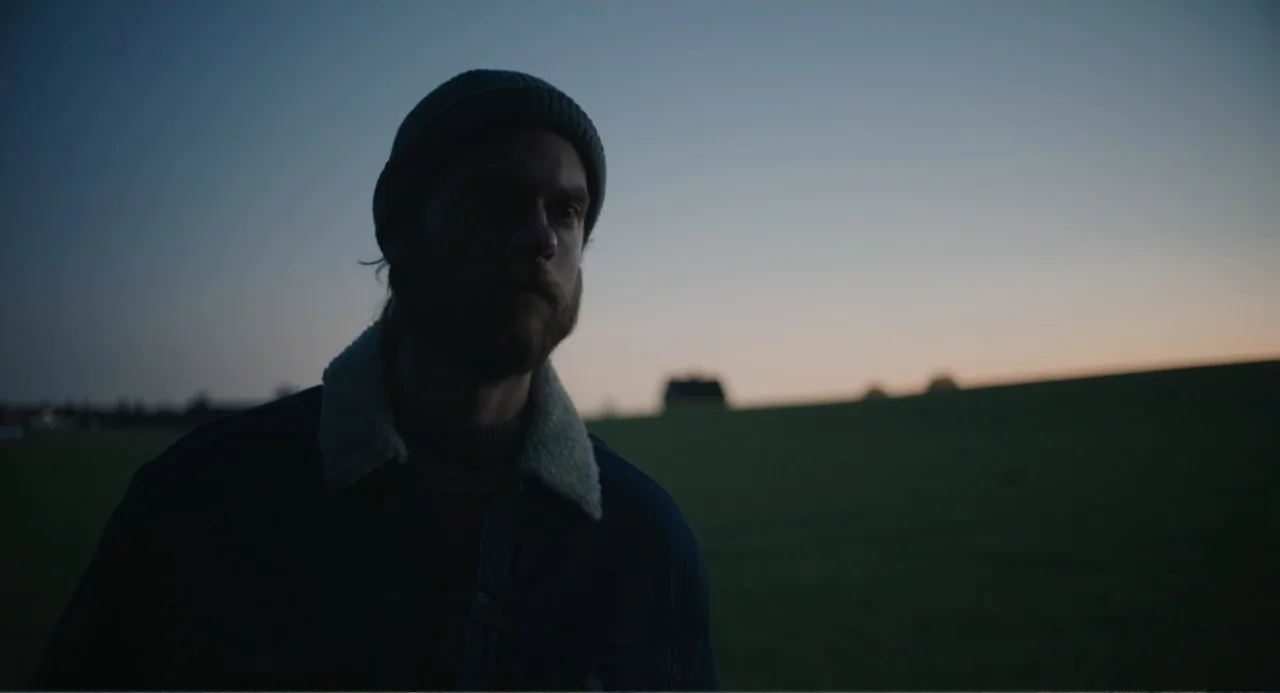 Ninjababy (2021) movie still: medium — A man stands in a dark, expansive open field at sunset under a dim sky. The subject wears a sherpa-collared…