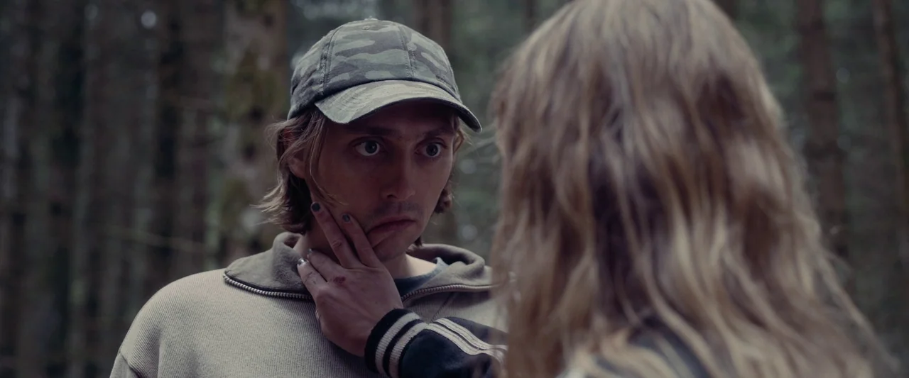 Meanwhile On Earth (2024) movie still: medium — A dense, dimly lit pine forest provides the background for an interaction between two people. A…