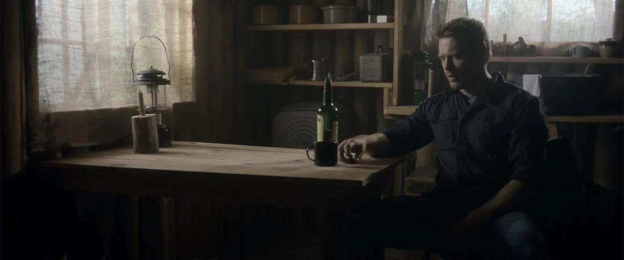 Radius (2017) movie still: medium — A rustic cabin interior features wood walls and shelves filled with basic kitchen supplies. A man in a dark…