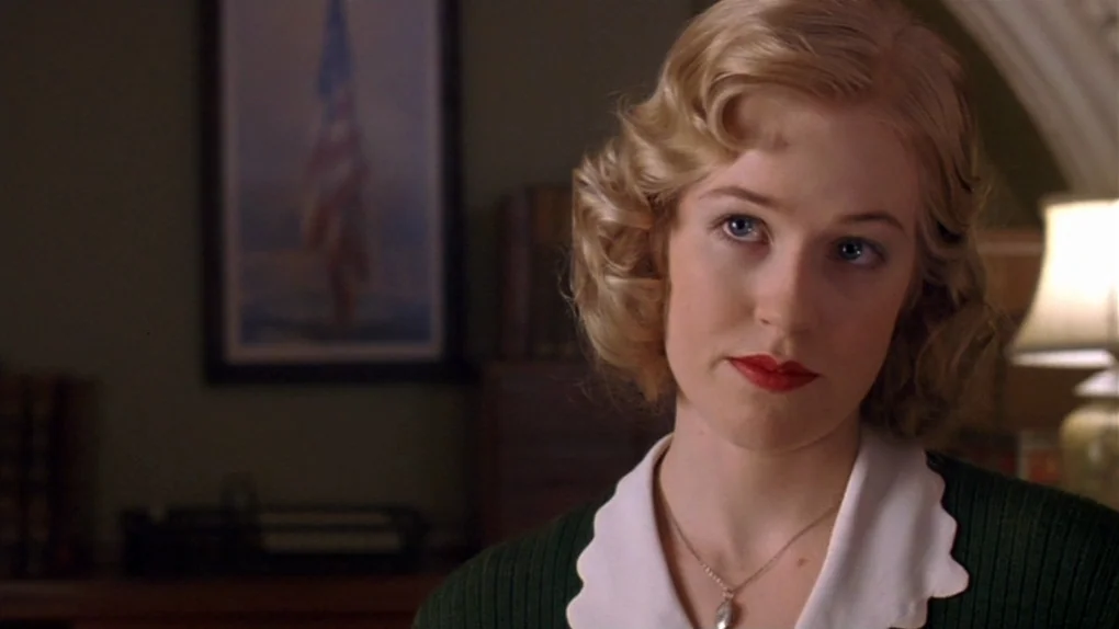 Ask The Dust (2006) movie still: closeup — A dark, wood-paneled office environment with a framed flag hanging on the wall. A woman with blonde wavy…