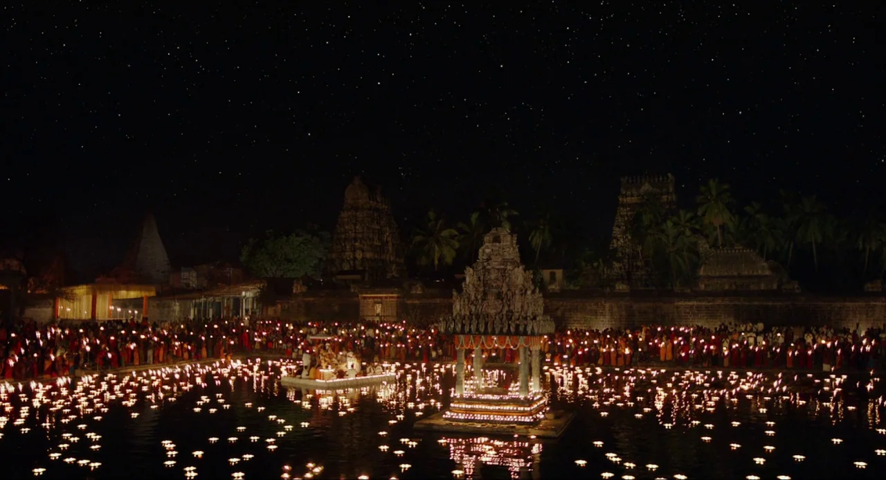 Life of Pi (2012) movie still: wide — A large crowd gathers around a stone temple tank at night under a starry sky. Thousands of small oil lamps…