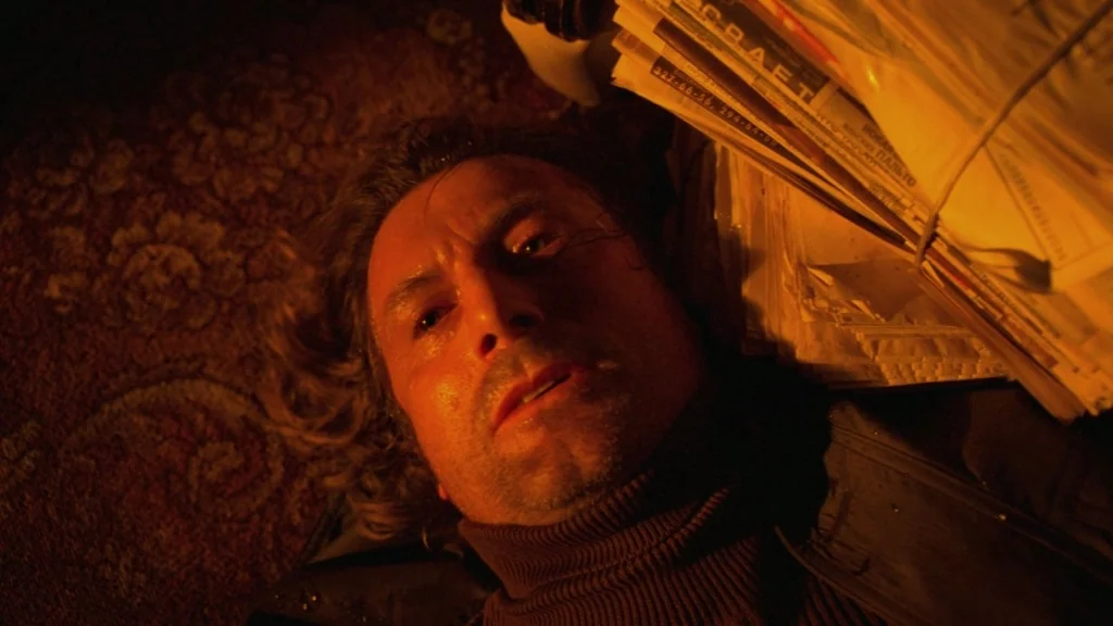 The Ugly Swans (2006) movie still: closeup — A man lies on a dark, intricately patterned carpet next to a stack of newspapers. The man looks upward…