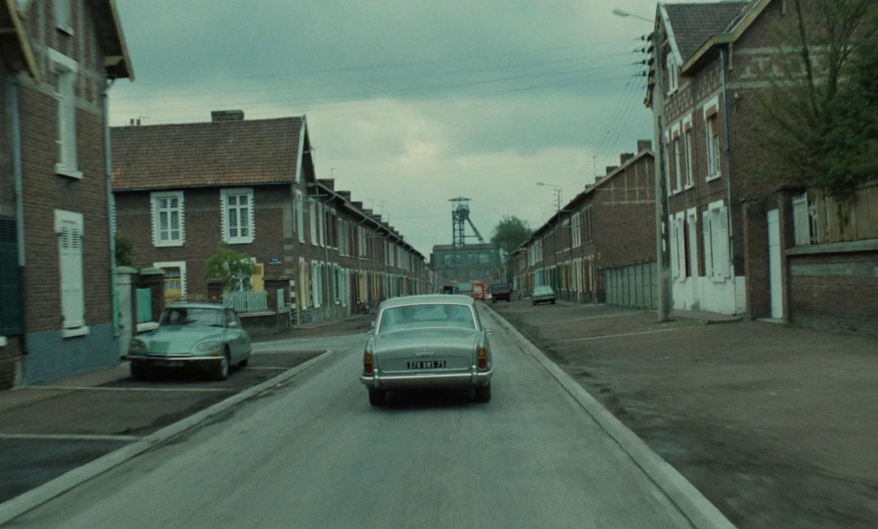 Graduate First (1978) movie still: wide — A quiet residential street in northern France features rows of uniform red-brick houses. A silver vintage…