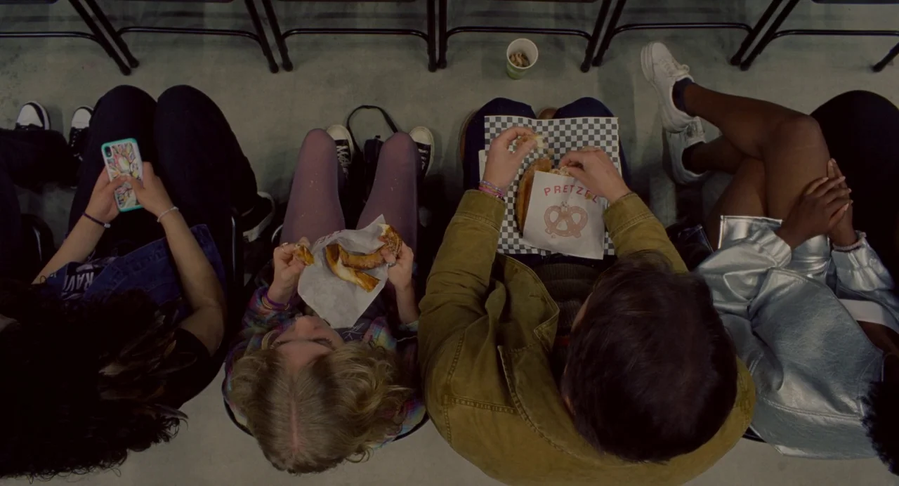 Trap (2024) movie still: wide — Four people sit in a row of folding chairs at an indoor event venue. They are eating soft pretzels from branded…