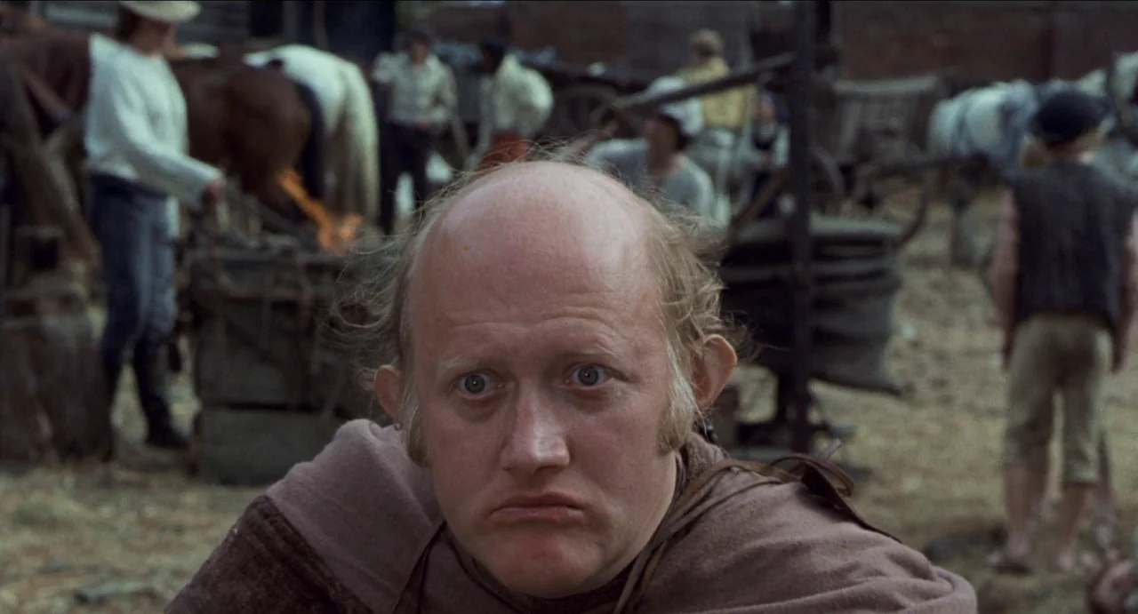 The Canterbury Tales (1972) movie still: closeup — A bustling outdoor medieval market with figures and animals in the blurred background. A balding…