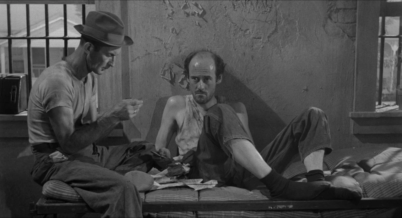 A Face In The Crowd (1957) movie still: medium — A spartan room with barred windows and peeling plaster walls suggests a confinement area. Two men…