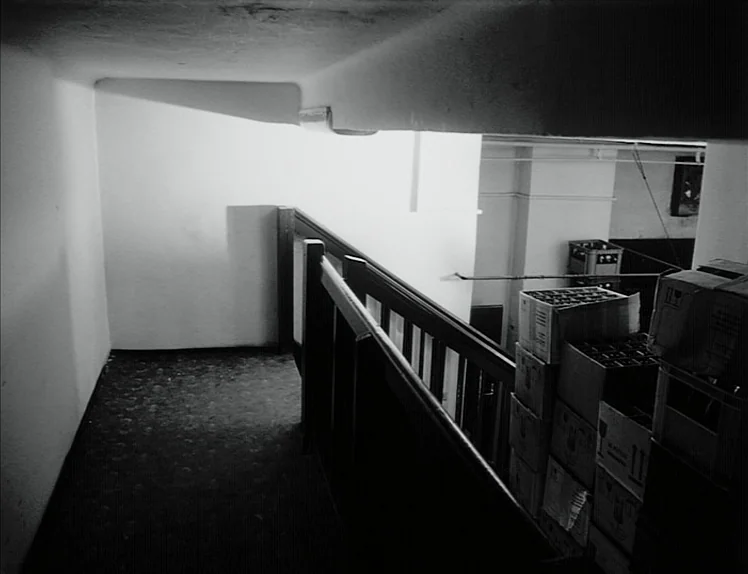 Damnation (1988) movie still: wide — A dimly lit industrial stairwell landing features stark white walls and a dark floor. Stacks of cardboard…