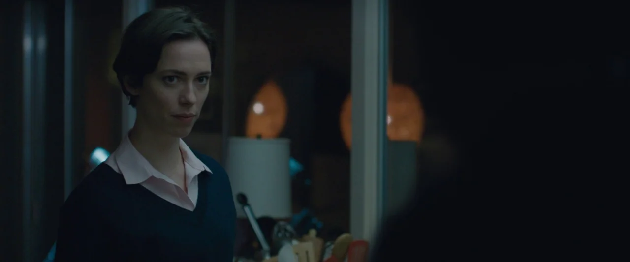 The Gift (2015) movie still: medium — A dimly lit interior space with a blurred kitchen counter and appliances visible. A woman stands in the…