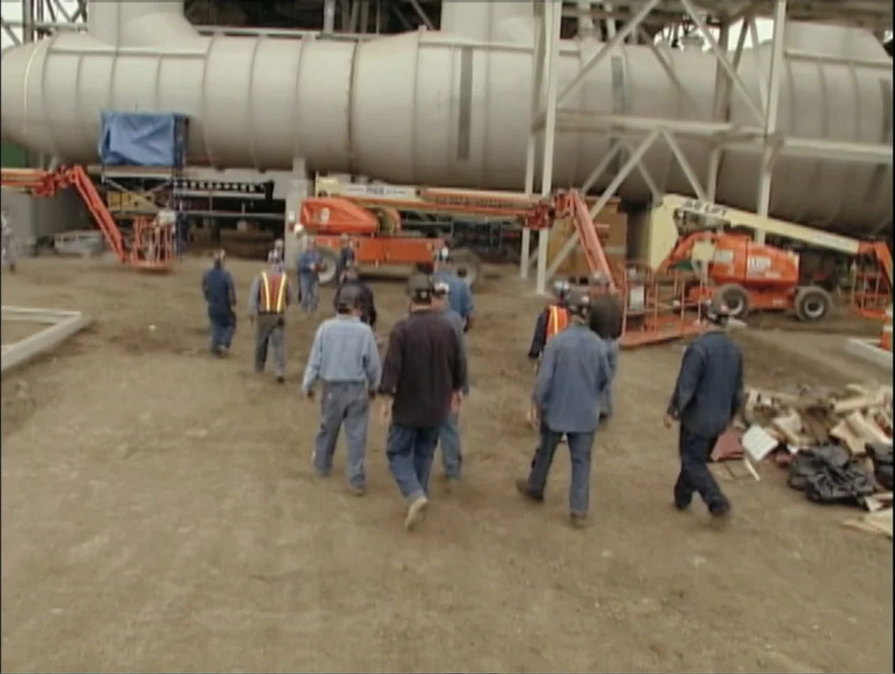 Addiction (2024) movie still: wide — A construction site features a large industrial pipe structure and orange mobile boom lifts. A group of workers…
