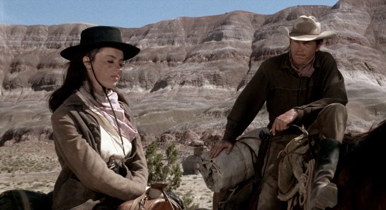 The Shooting (1966) movie still: medium — A vast arid desert with stratified, eroded rock formations fills the background behind riders. A man and a…
