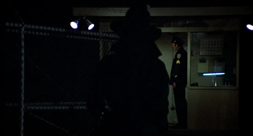 Family Plot (1976) movie still: medium — A night exterior showing a dark chain-link fence and a small illuminated guard shack. A figure in…