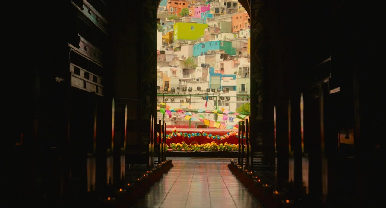 Diner (2019) movie still: wide — A dark, candle-lined interior aisle looks out through a stone archway at a town. The exterior reveals a hillside…