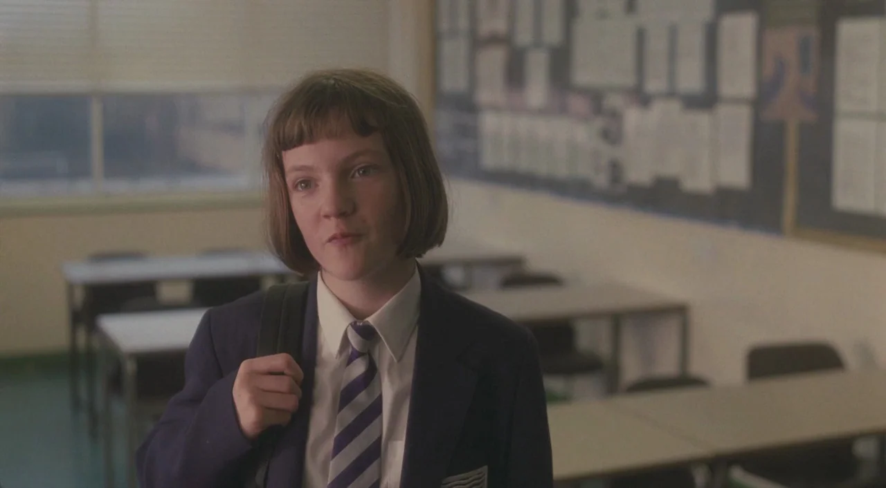 Broken (2012) movie still: medium — A quiet classroom contains rows of empty desks and a bulletin board covered in papers. A young girl in a school…
