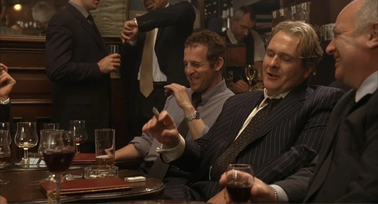 Scoop (2024) movie still: medium — A dark wood panelled pub interior with men seated at a circular table. Four men in suits talk and smoke cigars…