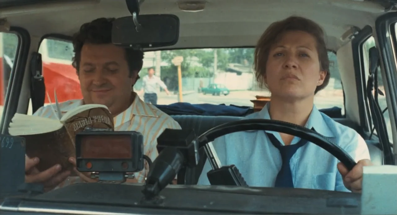 Do Not Expect Too Much from the End of the World (2023) movie still: medium — A man in a striped shirt and a woman in a blue uniform are in a car.…