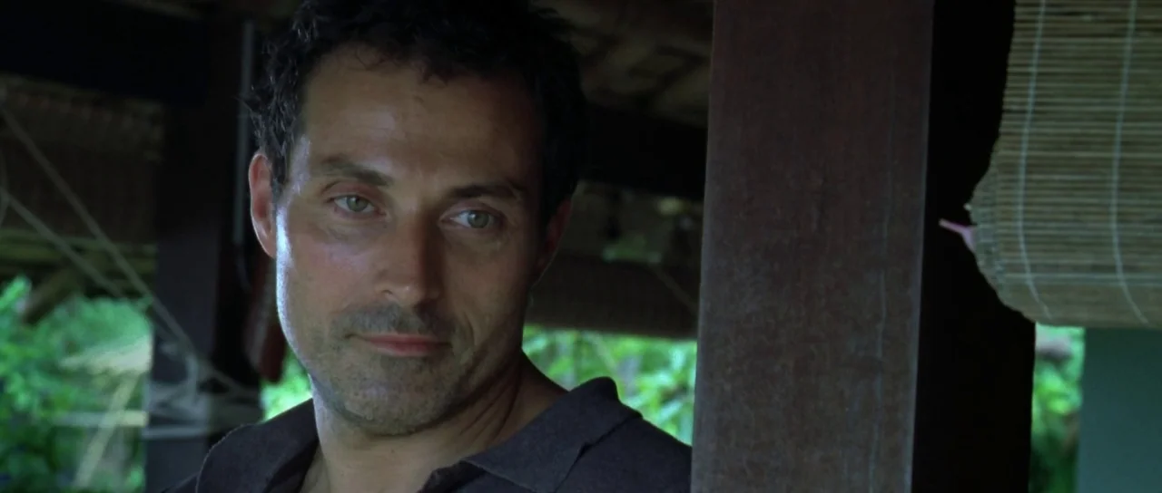 Vinyan (2008) movie still: closeup — A man stands on a wooden-pillared tropical porch with bamboo blinds in the background. He looks toward the side…