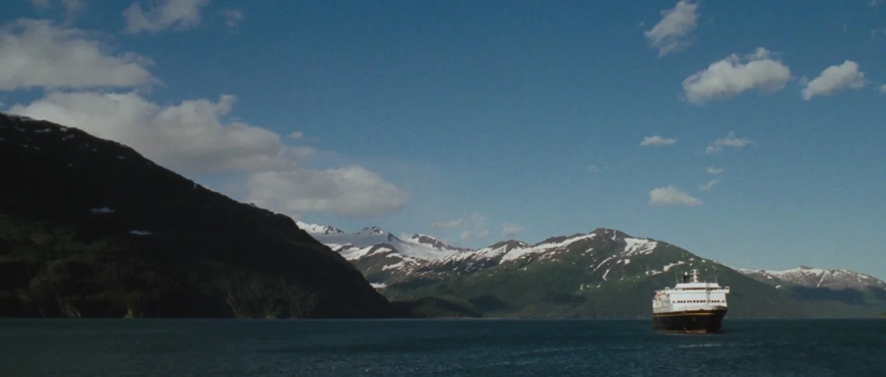 Wildlike (2014) movie still: establishing — A large ferry boat navigates a vast body of water surrounded by high mountains. The mountain slopes…