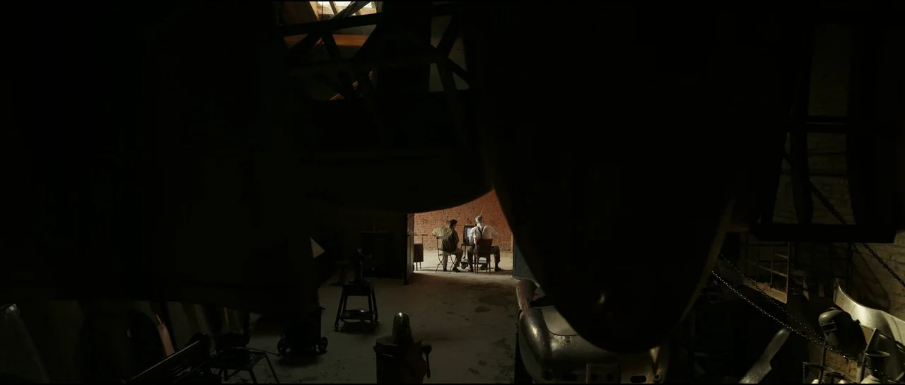Ferrari (2023) movie still: wide — A cluttered, dark industrial space features large hanging metal components in the foreground. Two men sit on…