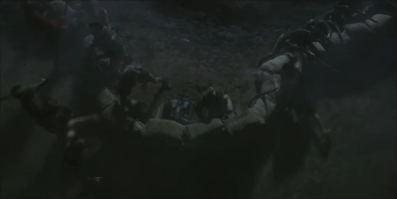 Exorcist: The Beginning (2004) movie still: wide — A dark muddy trench lined with stacked sandbags provides cover for soldiers. Several men crouch…