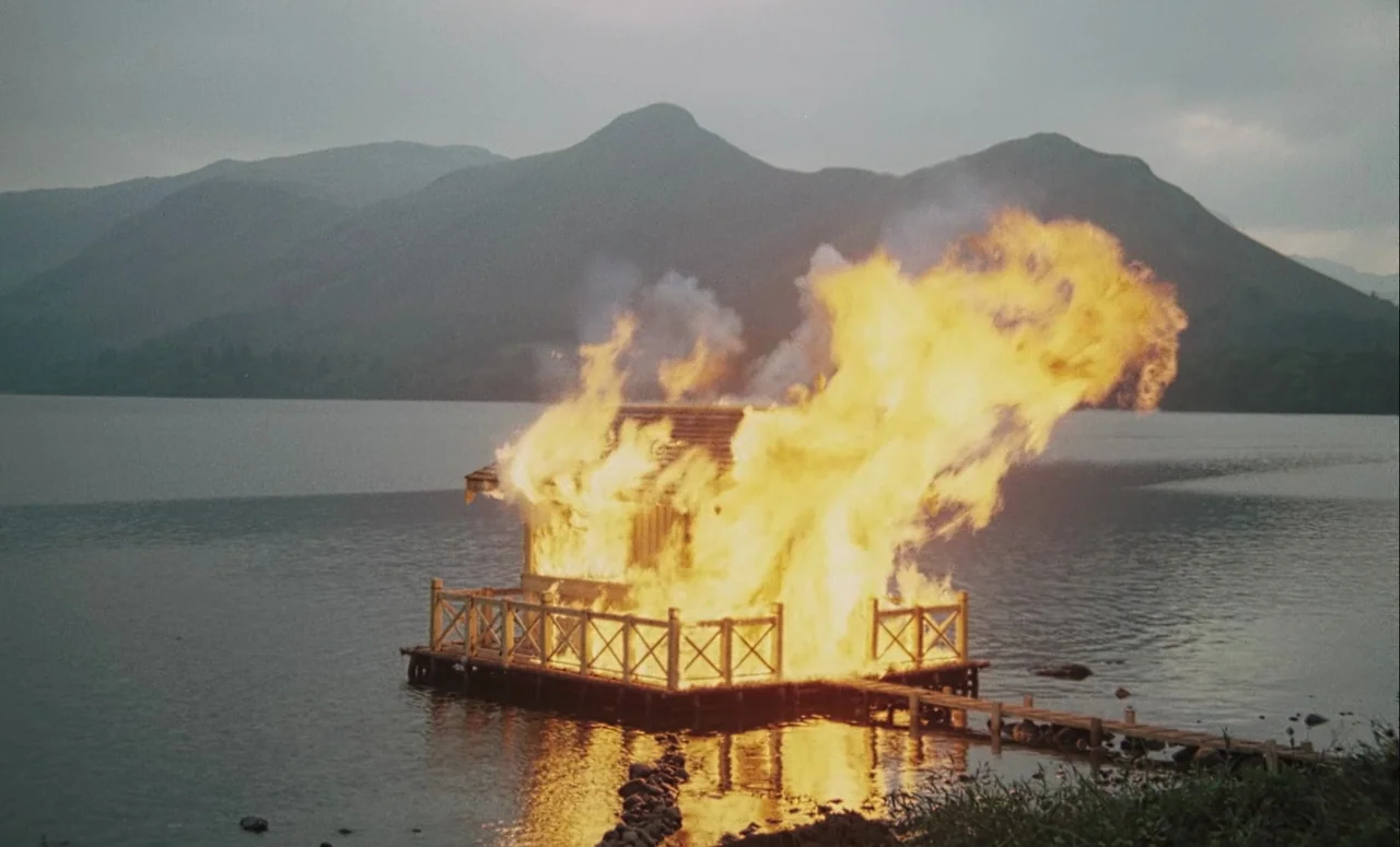 Mahler (1974) movie still: wide — A wooden structure on a platform in the middle of a lake is engulfed in flames. Large plumes of fire and smoke…