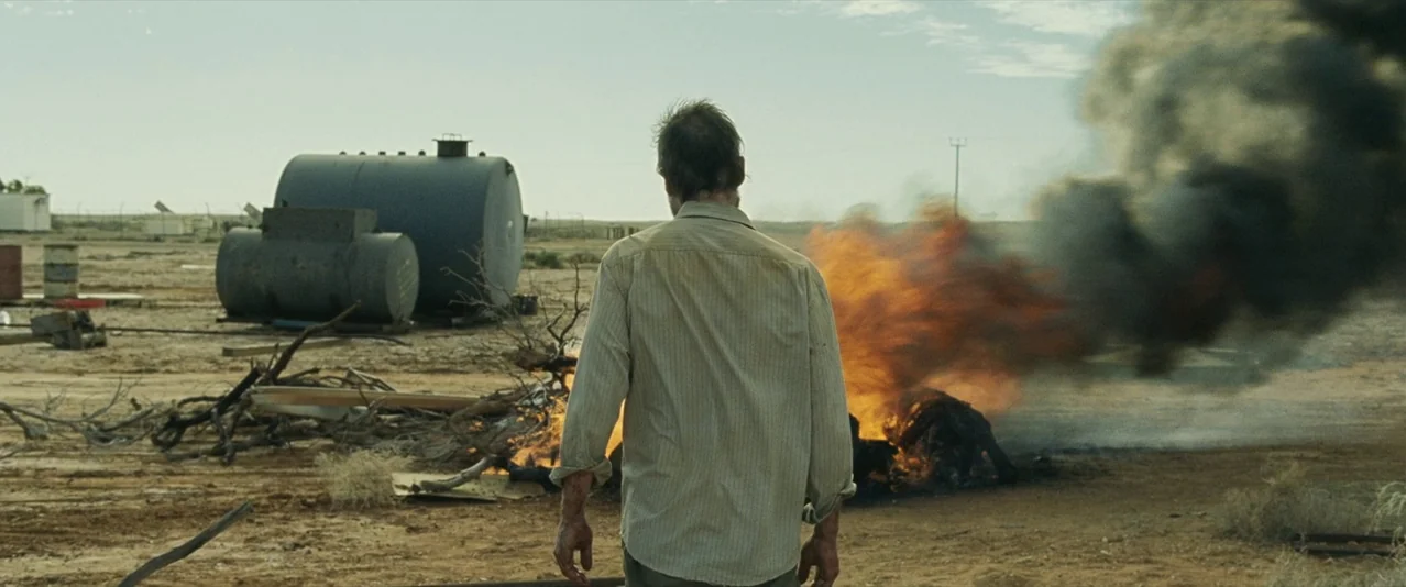 The Rover (2014) movie still: fullbody — A flat, arid landscape features industrial storage tanks and scattered debris under a blue sky. A lone man…